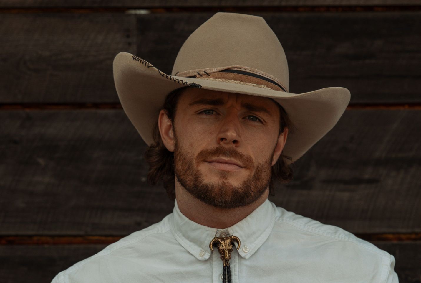 A man wearing a beige cowboy hat, white shirt, and gold bolo tie stands in front of some wooden paneling